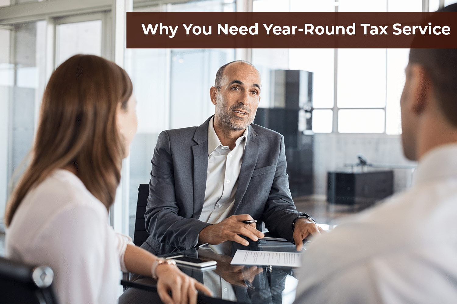 Two anonymous people gettin year round tax service advice from a bald man in a grey suit sitting across a glass table from them.