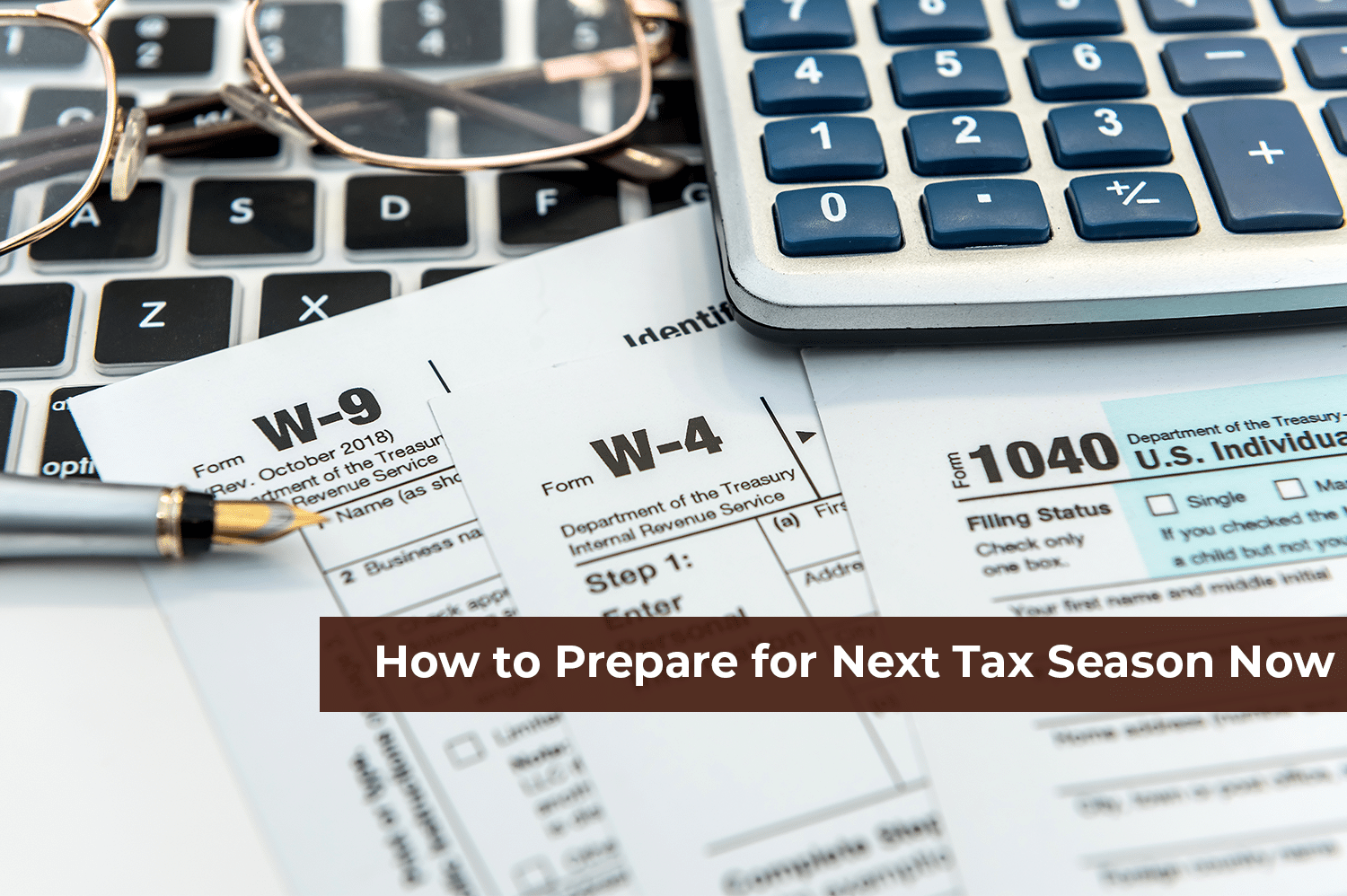 A close up of a 1040, W-4, and W-9 tax documents on a laptop computer with a pen, a pair of eye glasses, and a calculator laying on top, ready for next tax season.